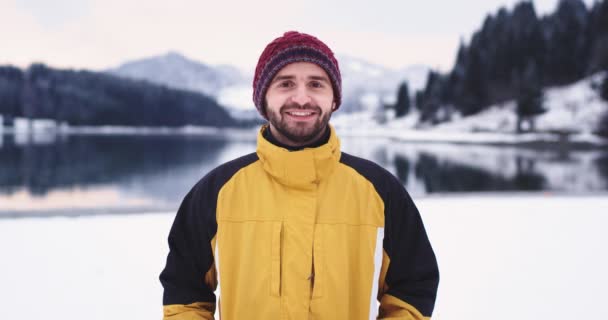 Ritratto di uomo carismatico in giacca arancione davanti alla telecamera sorridente grande qualcuno spara la palla di neve nel suo cappello momenti divertenti fuori in mezzo al paesaggio con lago e montagna innevata — Video Stock