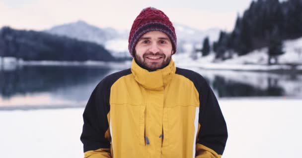 Πορτραίτο ενός άντρα στη μέση του τοπίου, κοιτάζοντας κατευθείαν στην κάμερα και χαμογελαστός μεγάλος πυροβολούσε από κάποιον με μια μεγάλη μπάλα χιονιού αστείες στιγμές το χειμώνα, φόντο εκπληκτικό — Αρχείο Βίντεο