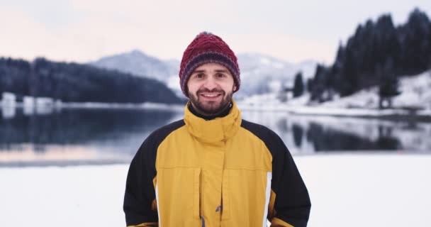 若いカリスマ的な男の肖像画は、カメラにまっすぐに見て、笑顔大彼は彼が美しい湖と山と奇跡的な場所で完璧な冬の日に雪の玉で撮影取得します — ストック動画