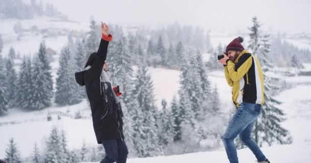 Двоє друзів щасливі приїхали в дивовижне місце, хлопець робить фотографії для свого друга в прекрасному місці в зимовий день, з засніженим лісом і горою навколо — стокове відео