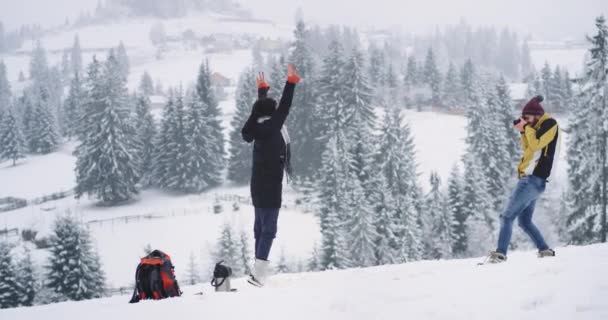 Dos turista llegó en lugar nevado increíble con hermoso bosque y montaña c hombre tomando fotos de su señora posando divertido delante de la cámara de fotos — Vídeo de stock