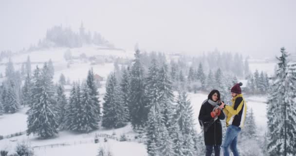 Jonge aantrekkelijke man en dame staan op de top van het veld achtergrond geweldig besneeuwd bos, ze kijken naar de foto 's van ze te nemen voordat — Stockvideo