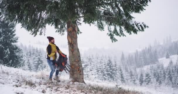 Alleinreisender Touristenmann im Winter inmitten der Berglandschaft erkundet er den Ort, hält unter einem großen Baum inne und setzt seine Fahrt durch das verschneite Feld fort — Stockvideo