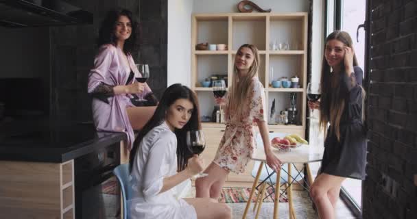 Багато етнічних жінок в дивовижній стильній піжамі насолоджуються часом разом в сучасному дизайні квартири, тримаючи келихи вина і дивлячись прямо на камеру . — стокове відео
