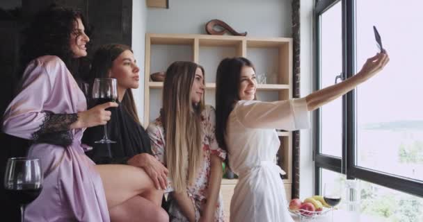 Привабливі багато етнічні дами в піжамі в студії міського дизайну знімають селфі-відео, використовуючи смартфон, тримаючи винні окуляри, які вони відчувають себе щасливими — стокове відео