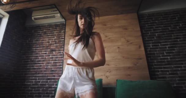 Mujer joven morena en pijama delante de la cámara saltando y bailando emocionada por la mañana en su moderno apartamento estudio — Vídeo de stock
