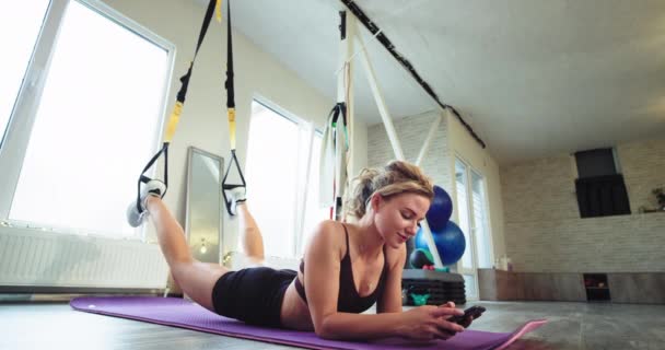 Stanco signora bionda dopo un esercizio di stretching corpo con elastici ha un tempo di relax chiacchierando sul suo smartphone mentre sdraiato sul tappeto sportivo — Video Stock