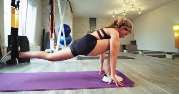 Концентрированная красивая женщина растягивает ноги, используя новые эластичные ленты, ремешки TRX, чтобы получить больше мышц в теле в большой просторной аэробной студии. 4k — стоковое видео