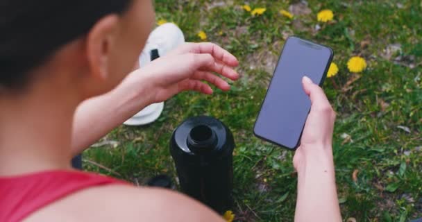 Details sportliche Dame in der Natur nach dem Training auf dem Boden sitzend und mit dem Smartphone etwas eintippen, was sie Pause macht, Wasserflasche hinter sich — Stockvideo