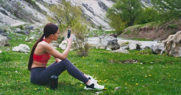 Mujer joven en una ropa deportiva disfrutando del tiempo después de un entrenamiento en la naturaleza se sentó en el suelo y tomar algunas fotos de hermoso paisaje a su alrededor — Vídeo de stock