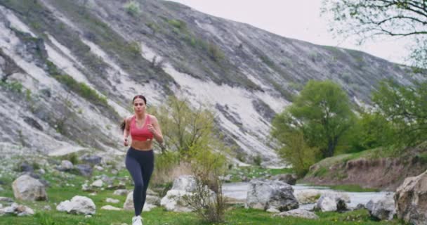 Hermosa dama con un cuerpo deportivo corriendo por el campo verde en un lugar hermoso increíble se cansa mientras hace su entrenamiento se detuvo y luego continuar su entrenamiento mientras escucha música usando un — Vídeos de Stock