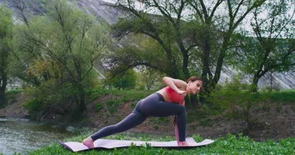 Nel mezzo di rocce e paesaggio fluviale giovane donna che pratica yoga sul tappeto lei rende il corpo allungamento posa felice lei godendo il momento in silenzio . — Video Stock