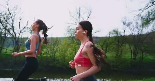 Two ladies with a fit bodies closeup running together in amazing landscape view wearing wireless airpods they enjoying the time together — Stock Video