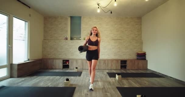 В удобной спортивной одежде после занятия йогой в студии велнес-йоги счастливая женщина со своим спортивным ковриком, идущая улыбаясь через студию перед камерой — стоковое видео