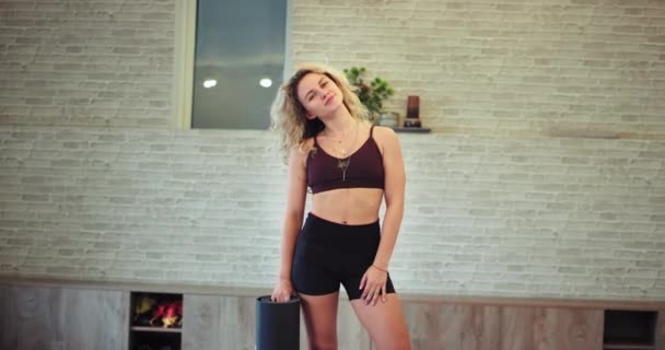 Харизматичная женщина с милой улыбкой в современной аэробной студии, стоящей перед камерой и держащей спортивный коврик, у нее крепкое мышечное тело — стоковое видео