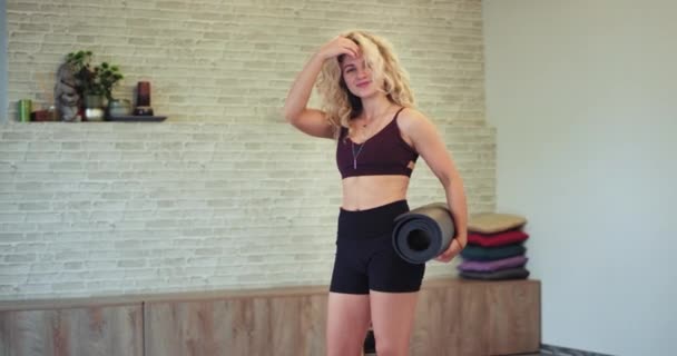 In een moderne aerobic Studio goed uitziende dame met een pasvorm lichaam op zoek rechtstreeks naar de camera charismatisch ze houden een sport mat en glimlachend schattig — Stockvideo