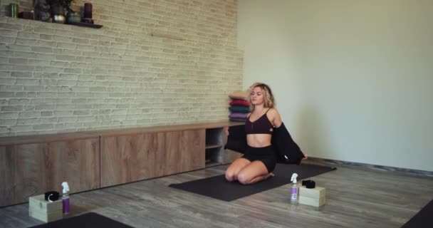 На спортивном коврике красавица, готовая к занятиям йогой в йога-студии, садится на пол — стоковое видео