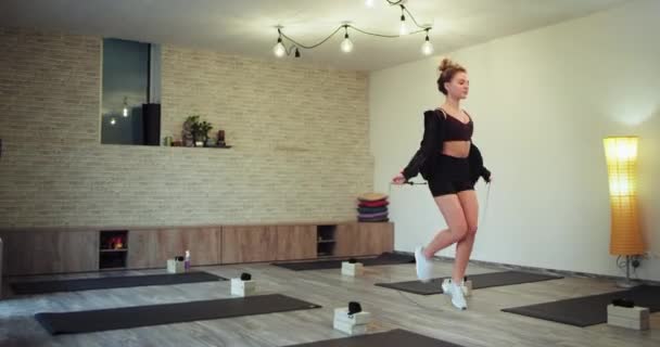 Mujer rubia carismática en ropa deportiva usando una cuerda para saltar para conseguir más músculo muy concentrado en un gran estudio aeróbico — Vídeo de stock