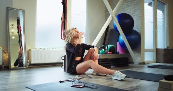 Señora sedienta después de un duro ejercicio de gimnasia que se sienta en el suelo bebiendo un poco de agua y neumático tener un tiempo de relax — Vídeos de Stock