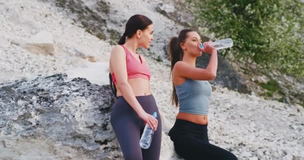 Zwei durstige Damen nach hartem Training in der Natur neben einem großen Stein stehen und Wasser aus der Plastikflasche trinken — Stockvideo