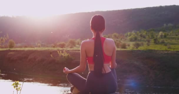 Młoda dama uprawiająca jogę na łonie natury w środku niesamowitego krajobrazu z rzeką ma medytację stwarza z powrotem do aparatu — Wideo stockowe