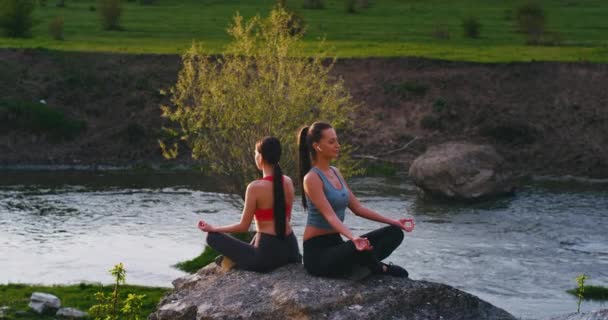 Dos amigas damas de la naturaleza en un lugar increíble con un lago practican yoga meditando posan sobre unas grandes piedras que se sienten relajadas y concentradas — Vídeo de stock