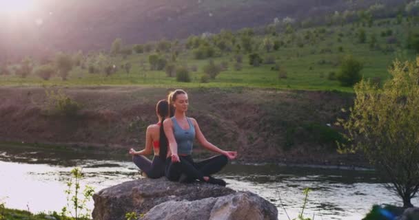 En una hermosa vista de la naturaleza con un lago y un campo verde, dos damas haciendo poses de meditación sobre grandes piedras se sienten concentradas y relajadas. — Vídeo de stock