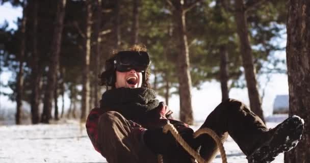 Chico muy feliz y sonriente usando unas gafas de realidad virtual mientras está sentado en el trineo en medio de la naturaleza juega un juego interesante muy entusiasta juega — Vídeos de Stock