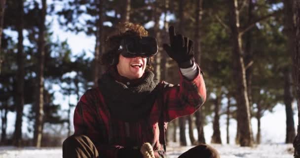 Porträt vor der Kamera charismatischer Mann mit lockigem Haar auf dem Schlitten mitten in der Natur an einem Wintertag mit einem Vr, während er auf dem Schlitten sitzt und virtuell mit dem Finger die — Stockvideo