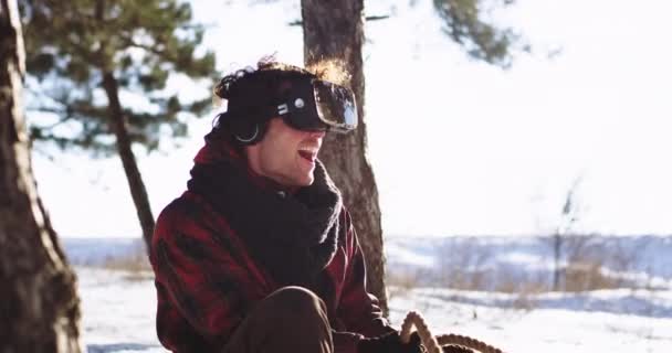 Chico guapo disfrutando del tiempo mientras está sentado en el trineo en medio del día de invierno utilizando unas gafas de realidad virtual para explorar la nueva generación de juego virtual muy impresionado de que jugar para — Vídeos de Stock