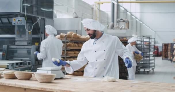 Aantrekkelijke en charismatische Bakker zeer stijlvolle dansen grappig in een bakkerij fabriek tijdens het vormen van stukjes deeg voor het bakken van brood, genietend van de tijd op zijn werkplek naast andere Baker het instellen van de — Stockvideo