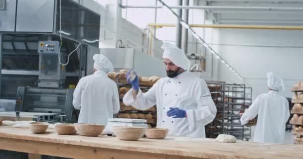 Σε μια βιομηχανία αρτοποιίας, ο χαρούμενος χορευτής φούρναρης διασκεδάζει ενώ ετοιμάζει τη ζύμη για να κάνει ένα ψωμί, μερικοί εργάτες να εργάζονται συγκεντρωμένοι πίσω του, το εργοστάσιο της βιομηχανίας τροφίμων — Αρχείο Βίντεο