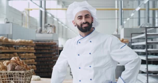 Na frente da câmera retrato de um homem bem parecido padeiro rosto comercial em um uniforme elegante sorrindo grande ao lado de uma mesa de trabalho em uma cozinha de padaria. 4k — Vídeo de Stock