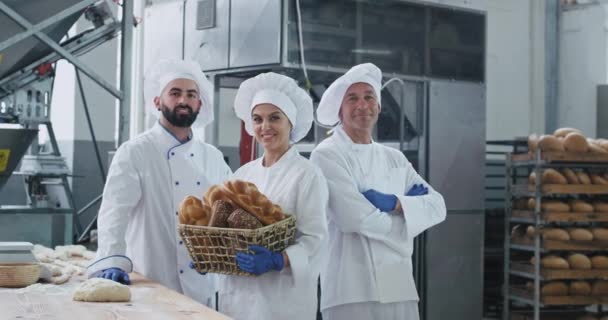 大きなパン屋業界で魅力的な3人のパン屋の魅力的な顔は、大きな笑顔で、オーガニックパンとヴィンテージバスケットを持つカメラにまっすぐに見えます — ストック動画