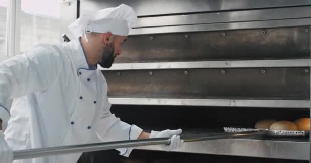 Endüstriyel fırın makinesinden pişmiş ekmek çıkarmak için özel bir meşale kullanarak sakallı karizmatik fırıncı — Stok video