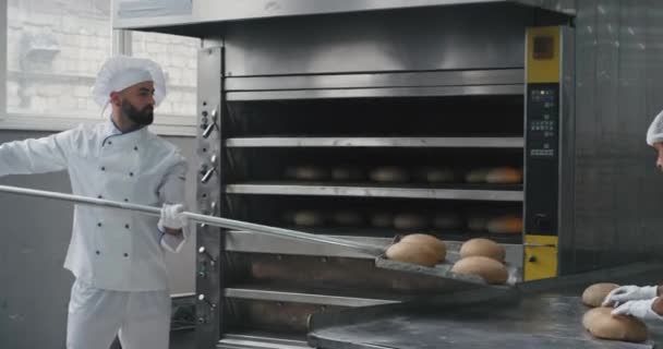 Panadería comercial panadero carismático y su asistencia profesional quitar el pan cocido de la máquina del horno y cargar el pan en los estantes — Vídeo de stock