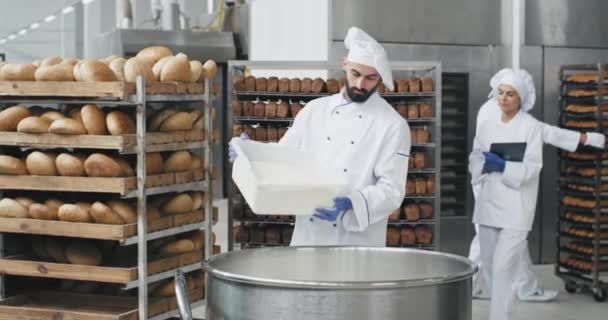 Przemysł piekarniczy duży szef kuchni w specjalnym mundurze przygotowującym ciasto dodać trochę mąki z koszyka piekarnika monitorowania wszystkich procesów tła wypiekany chleb na półkach. powolne ruchy — Wideo stockowe