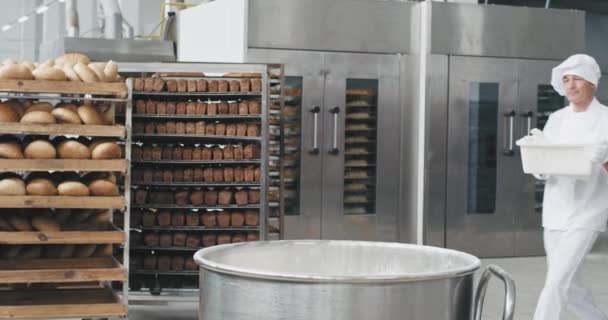 Bakkerij fabriek oude man chef-kok het deeg bereiden in een industriële container hij voegt wat meel toe terwijl op de achtergrond bakkerij medewerkers het gebakken brood vervoeren. 4k — Stockvideo