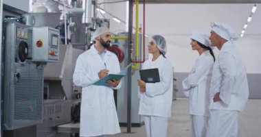 Yiyecek endüstrisi mühendisi elinde bir tablet tutuyor ve işçilerine makineyle ilgili talimatları anlatıyor. Korunmak için özel kıyafetler giyiyorlar.