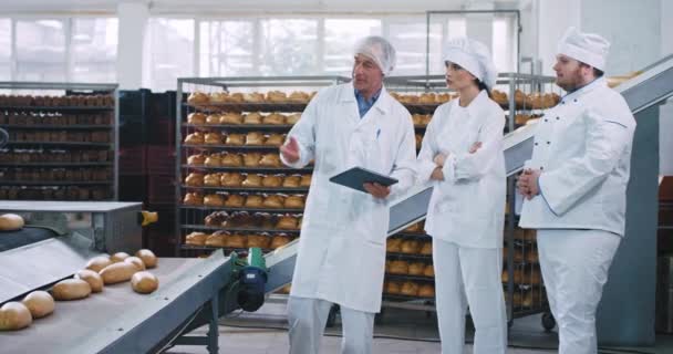 Alter Mann Ingenieur erklärt dem Bäckermeister Big Man und seiner Assistenz die Regeln für die Verwendung der automatischen Förderlinie, sie tragen spezielle Uniform — Stockvideo