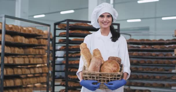 Señora panadero con una hermosa sonrisa sosteniendo una cesta con pan recién horneado y mirando directamente a la cámara en una industria de panadería — Vídeo de stock