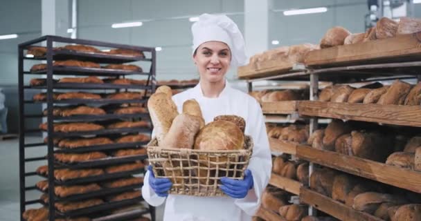 Retrato de mujer sonriente panadero con una cesta grande vintage con un pan fresco mirando directamente a la cámara y tienen una cara bastante comercial fondo grandes máquinas industriales modernas — Vídeo de stock