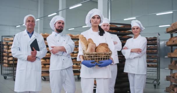 Bien parecido gran equipo principal de una fábrica de panadería sosteniendo una cesta con pan recién horneado mirando directamente a la cámara y sonreír lindo en un uniforme blanco — Vídeo de stock