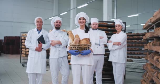Sonriente joven pastelera carismática y todo el equipo principal de panaderos en medio de la fabricación de panadería mirando directamente a la cámara y sonreír — Vídeo de stock