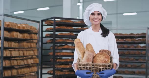 Gut aussehende junge Bäckerin hält einen frischen Vintage-Korb mit Bio-Brot und blickt direkt in die Kamera und lächelt sie trägt stylische Bäckeruniform — Stockvideo