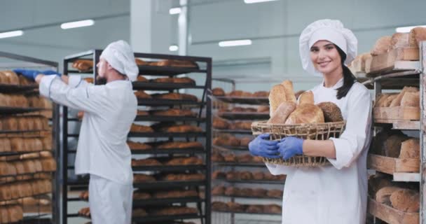 Attractive lady baker with a big basket of a fresh organic bread in bakery looking straight to the camera portrait smiling large background full shelves of bread other baker put them in order — Stock Video