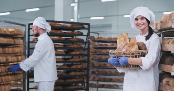 Lächelnd schöne junge Bäckerin mit einem Korb mit frisch gebackenem Brot schaut direkt in die Kamera und lächelt großen Hintergrund andere Bäcker Mann ordnen das Brot im Regal — Stockvideo