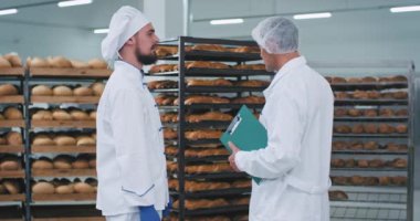 Genç fırıncı adam ve ana mühendis taze pişmiş ekmek mühendisi ile raflar çok bir fırın bölümünde bir konuşma var onlar özel beyaz üniforma giyen fırıncı bir şey açıklamak