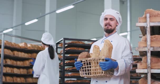 Добре виглядає чоловік з бородою і вусами пекар тримає великий кошик свіжого запеченого хліба, дивлячись прямо на камеру і посміхаючись, в той час як інша пекарня на фоні організовує хліб в порядку — стокове відео