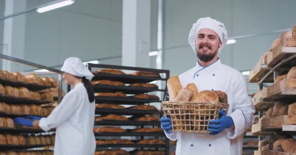 Charismatický usměvavý muž s bradou, držící košík plný čerstvě upečeného chleba a dívá se přímo do kamery v pekárenské branži další pracovník, který si uložil čerstvý pečený chléb — Stock video